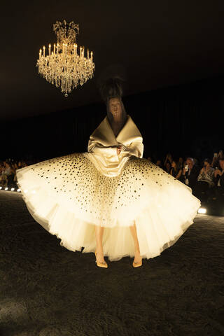 Model spotlit on a dark catwalk wearing a large white tulle gown with a chandelier above her
