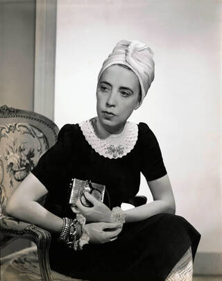 Black and white photograph of a seated woman wearing a black dress, white turban and holding a small purse