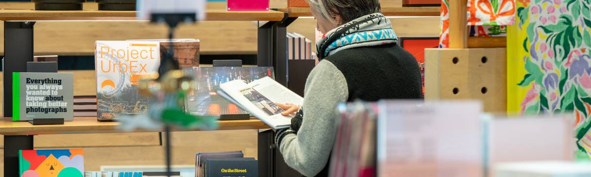 A person standing reading a book beside a bookcase.
